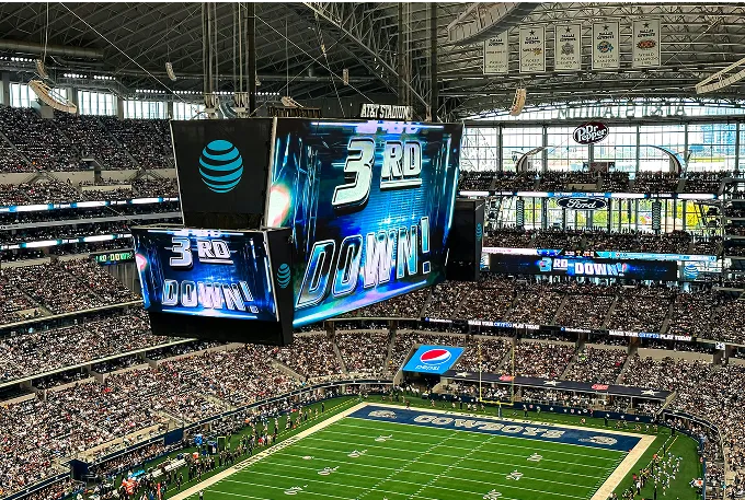 A large stadium filled with fans, featuring a prominent screen displaying an exciting football game.